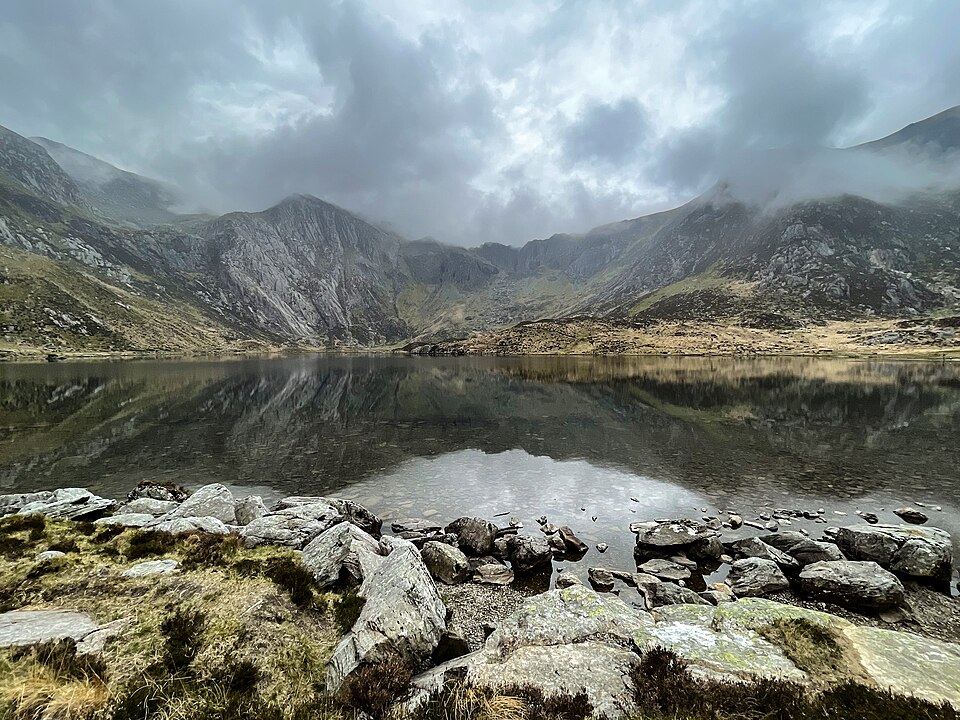 Photo of a lake in Cwm Idwal at the Glyderau mountain range. The sky is a cloudy grey, reflected in the glassy surface of the lake, which is surrounded by mountains in the background and lichen spotted rocks in the foreground.