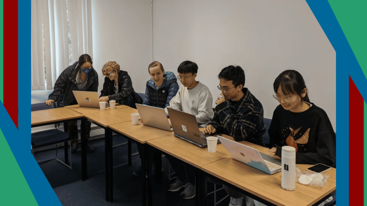 A photograph of a group of university students sat at a desk working on their laptops, framed by the Wikimedia UK colours of green, red and blue.